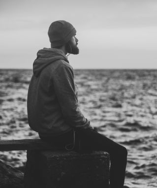 Grayscale Photo of Man in Hoodie and Kit Cap Sitting Near Bodies of Water